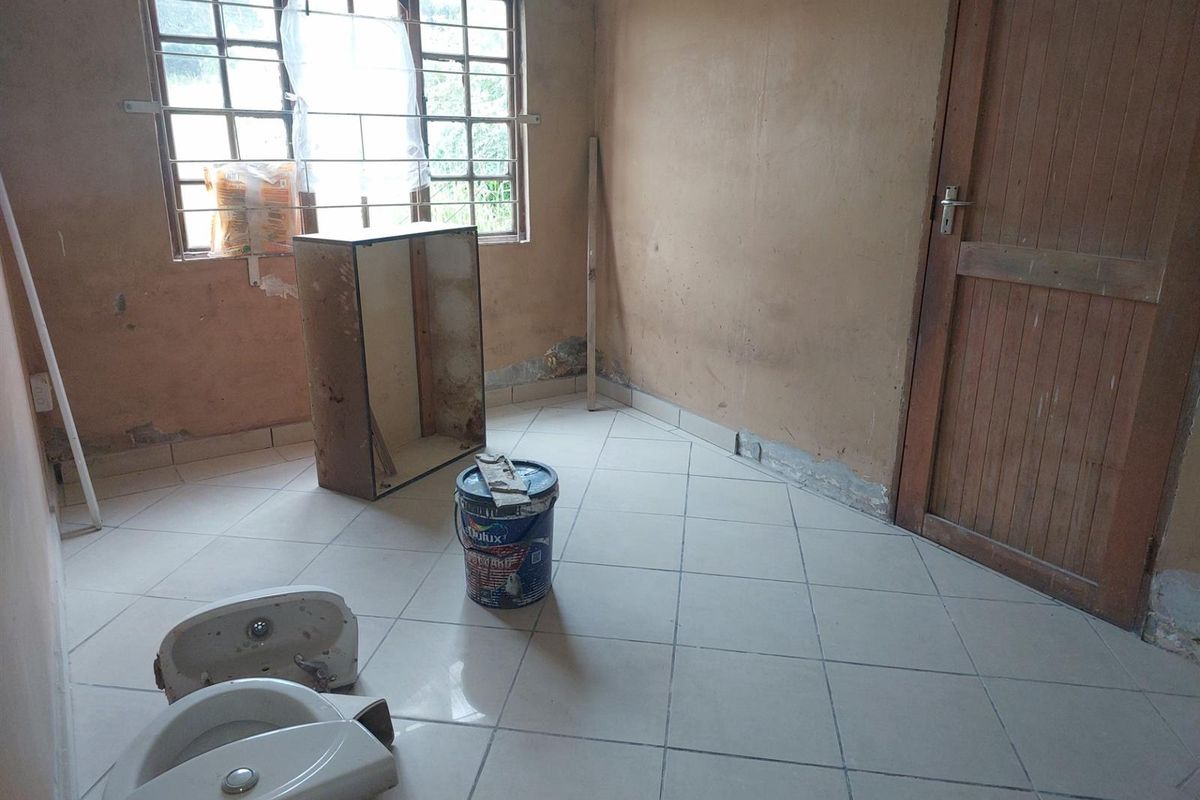 A vacant room undergoing renovation, featuring tiled flooring, a wooden door, and a window with security bars, currently in need of significant cosmetic repair.