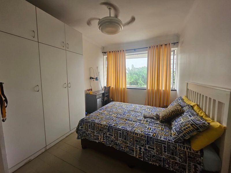 A functional bedroom featuring built-in cupboards, a ceiling fan, and window security bars, offering a practical layout with natural light.