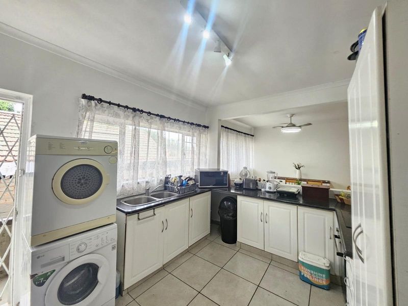 Functional kitchen featuring white cabinetry, dark countertops, double sink, and tiled flooring, with integrated laundry appliances and ample natural light.
