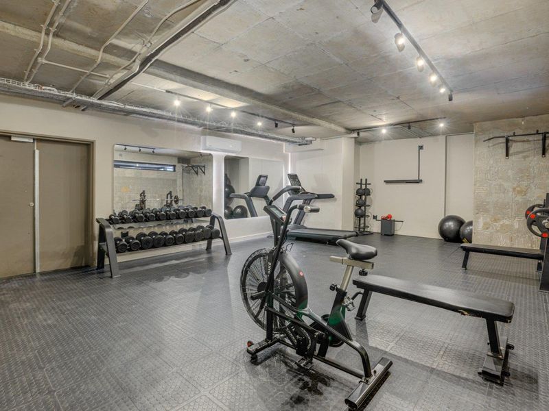 A well-equipped residential gym featuring industrial-style exposed concrete ceilings, track lighting, durable rubber flooring, and wall-mounted mirrors.