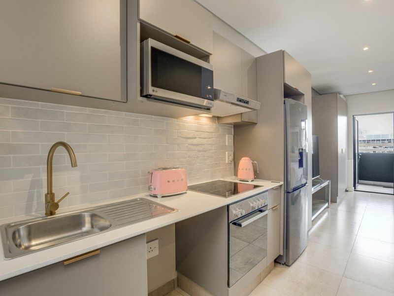 Modern open-plan kitchen featuring sleek grey cabinetry, stone countertops, brass fixtures, and integrated appliances, flowing into a bright living area with balcony access.