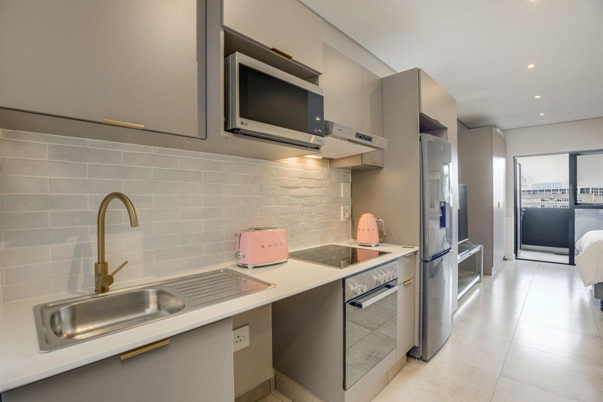 Modern open-plan kitchen featuring sleek grey cabinetry, stone countertops, brass fixtures, and integrated appliances, flowing into a bright living area with balcony access.