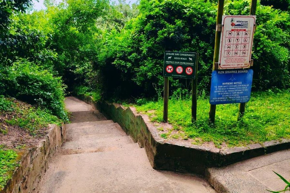 A concrete pathway leading through a lush, green garden area with signage indicating access to the beach.