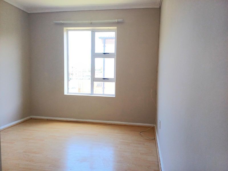 A bright, empty bedroom featuring light wood-look laminate flooring, neutral wall paint, and a large window with a curtain rail.