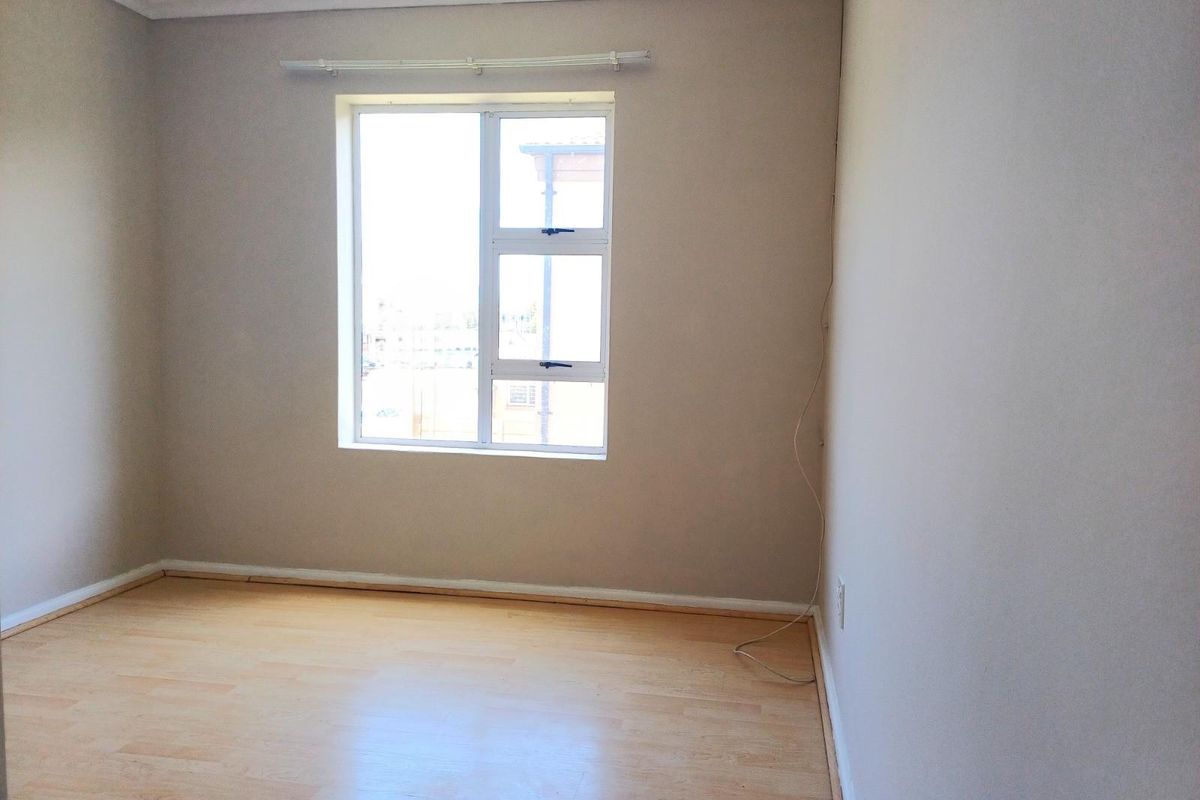 A bright, empty bedroom featuring light wood-look laminate flooring, neutral wall paint, and a large window with a curtain rail.