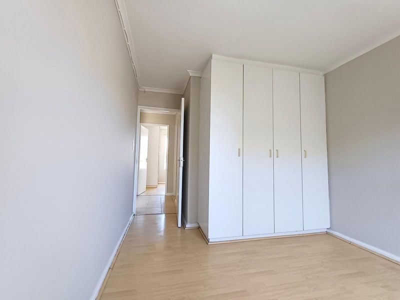 A bright, empty bedroom featuring light wood-look flooring, neutral walls, and a large, white built-in wardrobe with ample storage space.