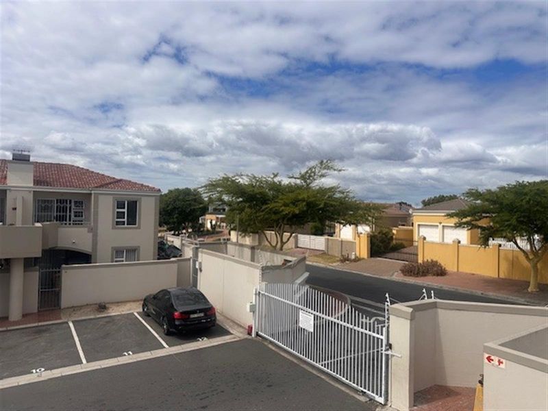 An elevated view of the apartment complex entrance, featuring paved parking bays, a secure gated access point, and surrounding residential boundary walls.