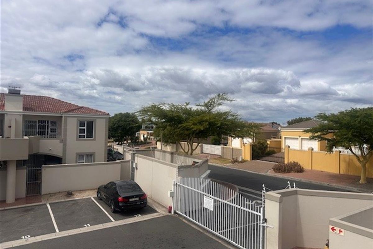 An elevated view of the apartment complex entrance, featuring paved parking bays, a secure gated access point, and surrounding residential boundary walls.