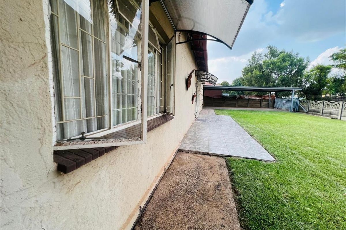 Rear exterior view featuring a tiled patio area, metal window awnings, and a spacious lawn with a carport and boundary wall.
