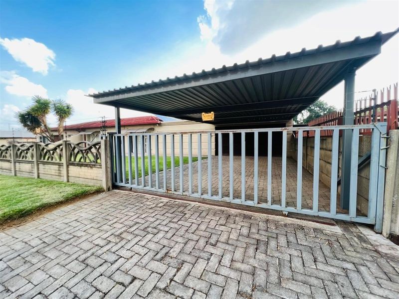 Front exterior view featuring a paved driveway, secure sliding gate, and a covered carport structure.