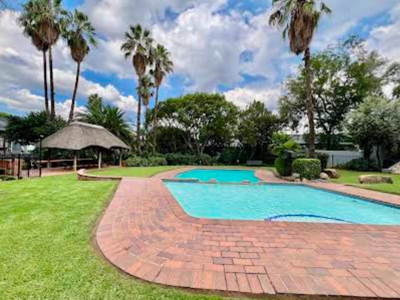 A well-maintained communal pool area featuring brick paving, a thatched lapa for outdoor entertaining, and established garden landscaping.