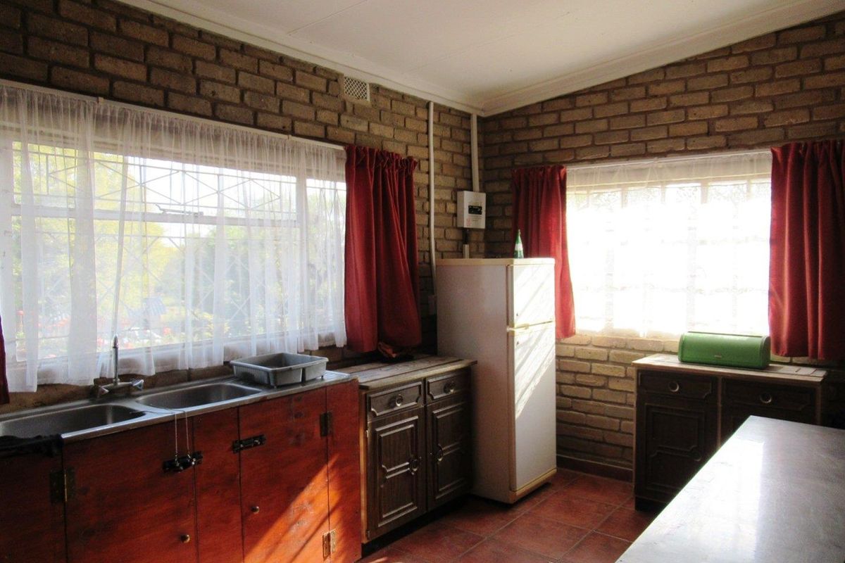 A rustic kitchen featuring exposed brick walls, tiled flooring, and wooden cabinetry with a double sink and large windows providing natural light.