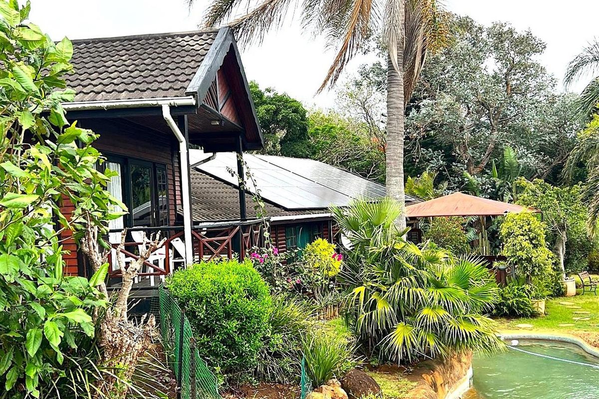 Rear view of a timber-clad farm dwelling featuring a raised deck, solar panel installation, and an adjacent swimming pool set within a lush garden.