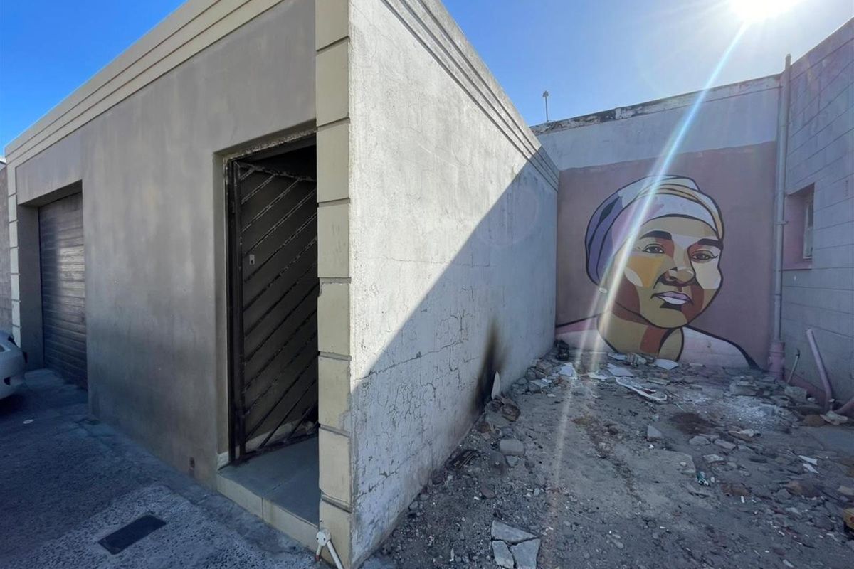 Rear exterior view of a property showing a security gate, concrete structure, and a mural on the adjacent wall with visible debris and surface damage.