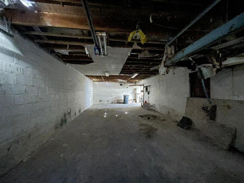 An unfinished, vacant garage space featuring exposed ceiling joists, concrete block walls, and a concrete floor, requiring significant renovation and repair.
