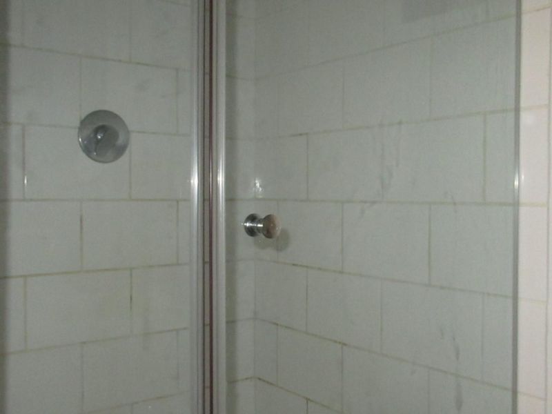 A compact shower enclosure featuring white wall tiles and a chrome mixer, requiring maintenance to address grout discoloration and surface wear.