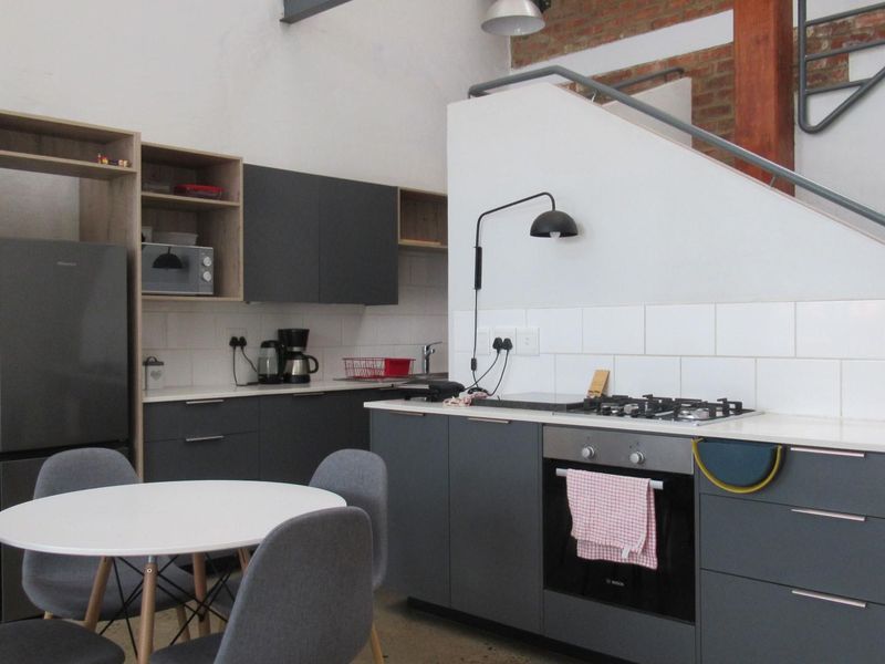 Modern open-plan kitchen featuring dark grey cabinetry, gas hob, built-in oven, and subway tile backsplash, set against an exposed brick wall and staircase.