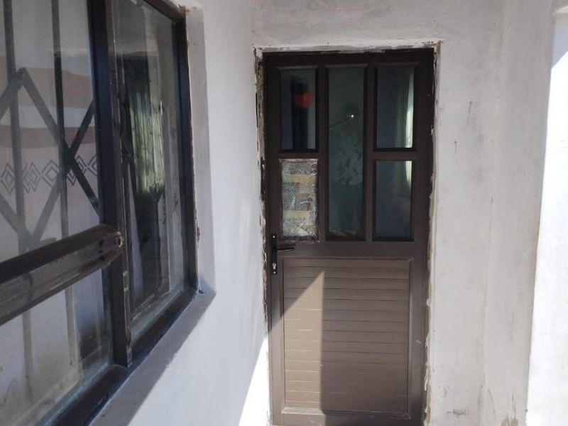 A rear exterior entrance area featuring a brown aluminium door and window, showing significant signs of wear and unfinished plasterwork around the door frame.
