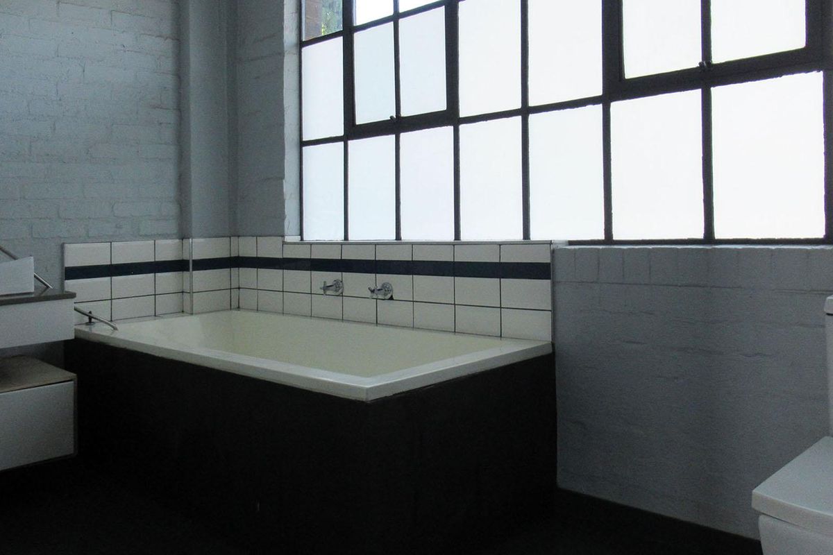 A spacious industrial-style bathroom featuring a built-in bathtub, wall-mounted basin, toilet, and large steel-framed windows with painted brick walls.