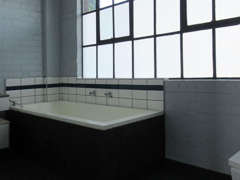 A spacious industrial-style bathroom featuring a built-in bathtub, wall-mounted basin, toilet, and large steel-framed windows with painted brick walls.