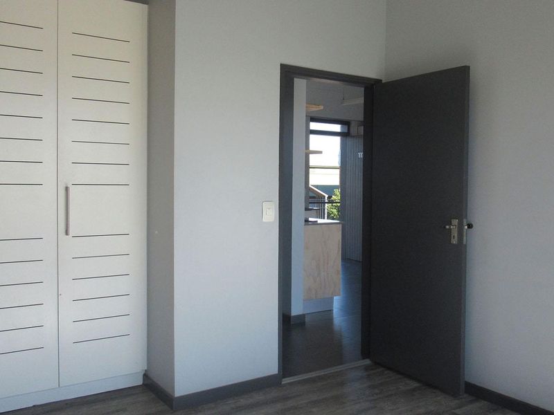 A clean, empty bedroom featuring a modern built-in wardrobe and neutral finishes, with a view into the adjacent living area.