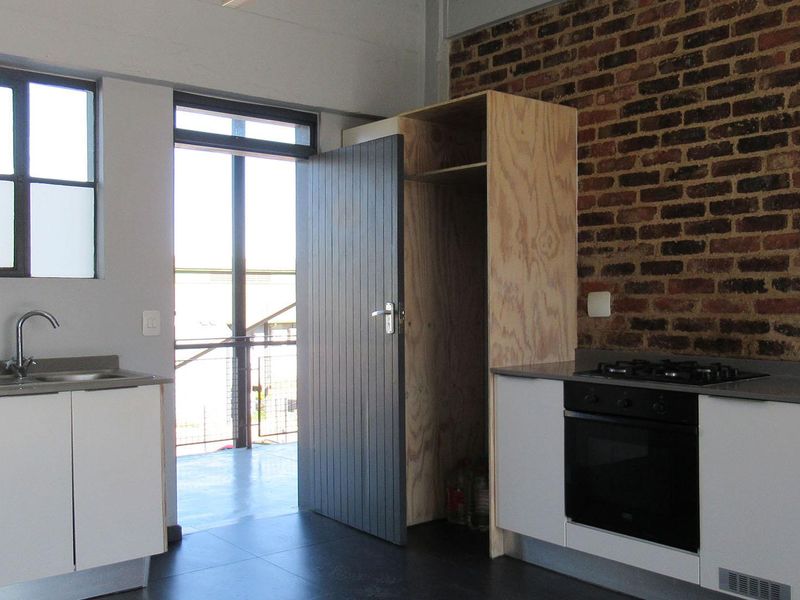 Compact kitchen featuring exposed brick walls, modern built-in oven and hob, stainless steel sink, and minimalist plywood cabinetry with dark tiled flooring.