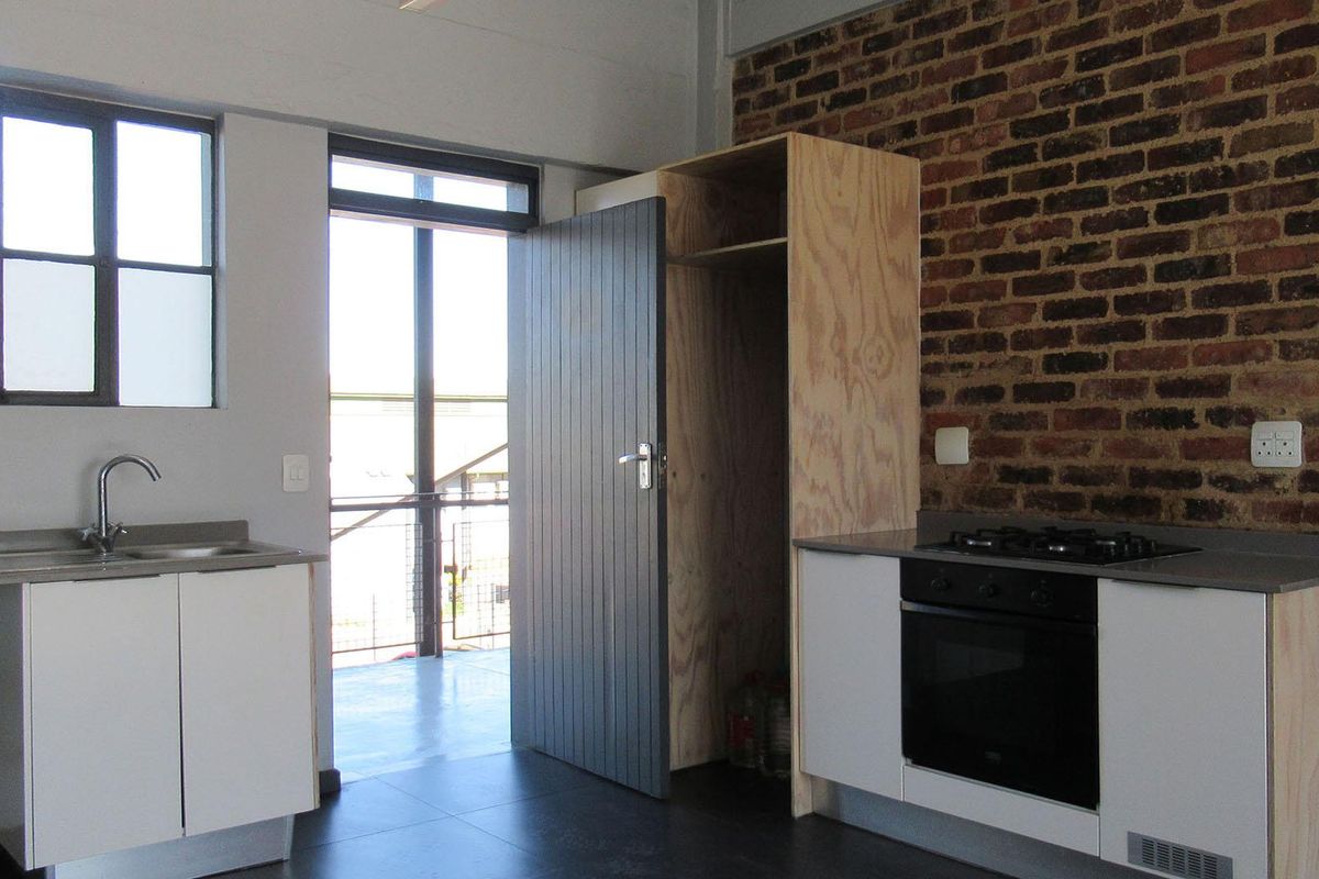 Compact kitchen featuring exposed brick walls, modern built-in oven and hob, stainless steel sink, and minimalist plywood cabinetry with dark tiled flooring.