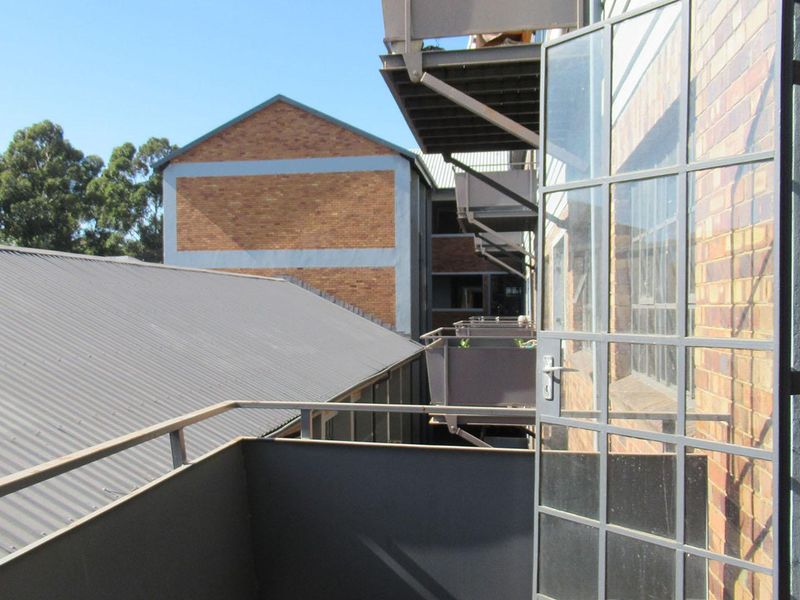A functional balcony with metal railings and a steel-framed glass door, overlooking adjacent industrial-style apartment buildings with corrugated metal roofing.