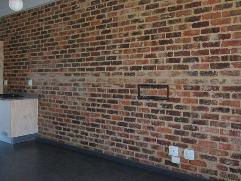 Open plan living area featuring a prominent exposed brick feature wall, tiled flooring, and a compact kitchen unit with built-in oven and gas hob.