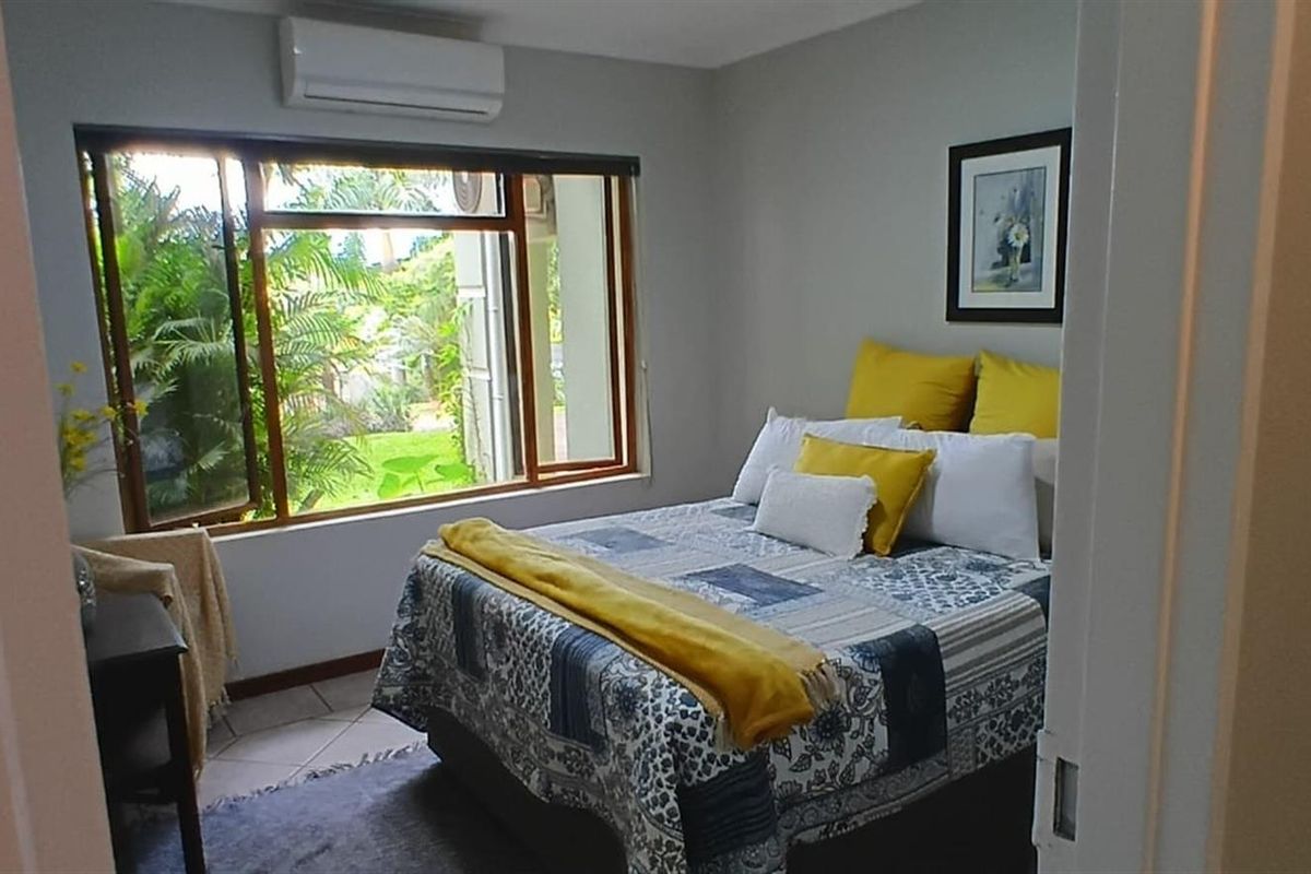 A well-lit bedroom featuring tiled flooring, a large window with garden views, and a wall-mounted air conditioning unit.