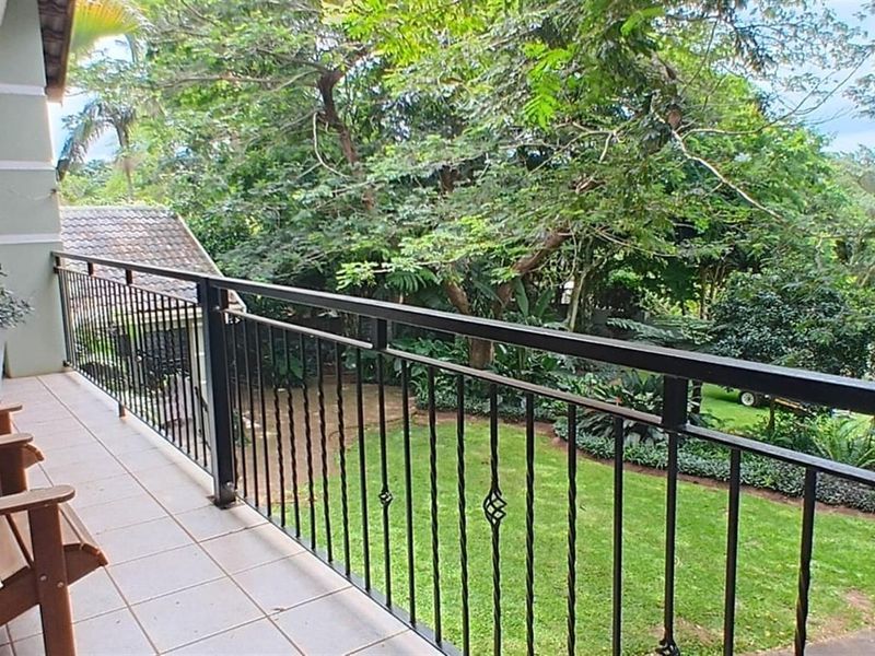 A covered balcony featuring tiled flooring and a black metal balustrade, overlooking a lush green garden and mature trees.