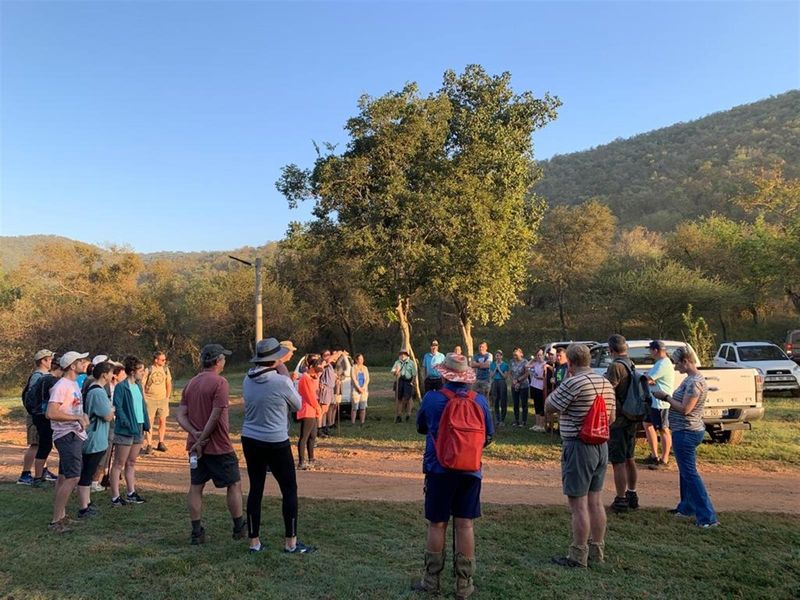 A group of people gathered on a dirt track in a natural, undeveloped outdoor setting with trees and hills in the background.