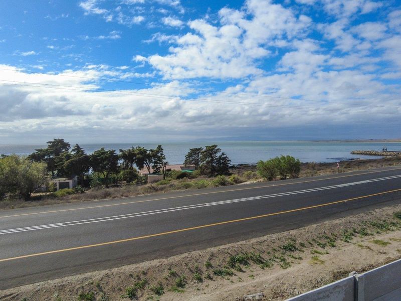 A scenic coastal view overlooking a public road, surrounding vegetation, and the ocean horizon under a partly cloudy sky.