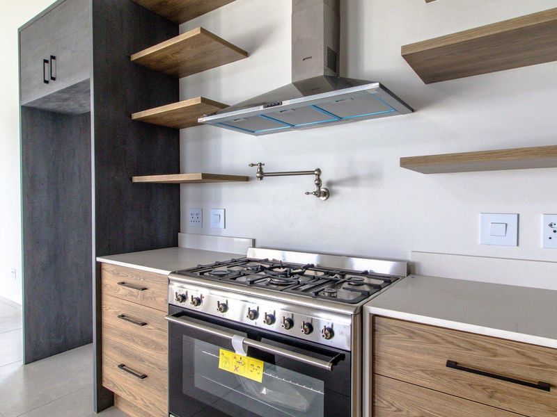 Modern kitchen featuring a gas stove, stainless steel extractor fan, pot filler faucet, floating shelves, and sleek wood-finish cabinetry with stone countertops.