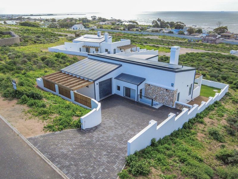 Aerial view of a modern coastal residence featuring a paved driveway, stone-clad facade, timber pergola, and solar panels, set in a scenic landscape.
