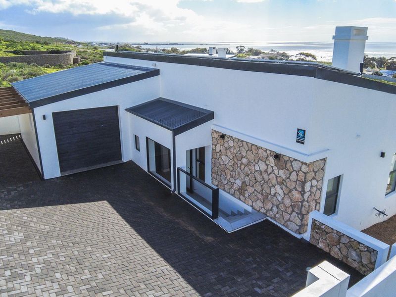 Aerial view of a modern coastal home featuring stone cladding, a paved driveway, and a garage, set against a scenic ocean backdrop.