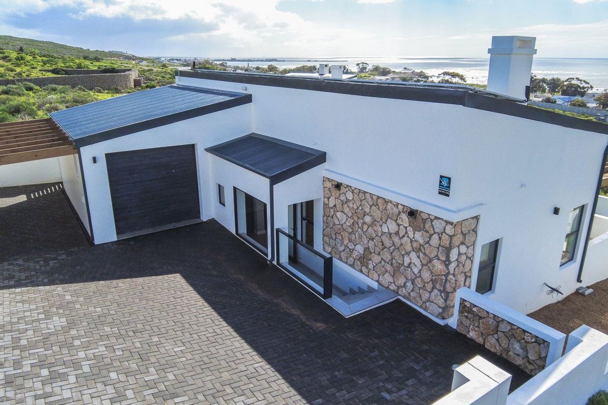 Aerial view of a modern coastal home featuring stone cladding, a paved driveway, and a garage, set against a scenic ocean backdrop.