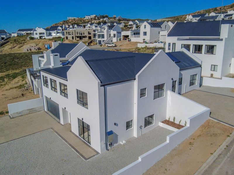 Aerial view of a modern white house with a dark metal roof, featuring a paved driveway and surrounding boundary walls in a coastal estate.