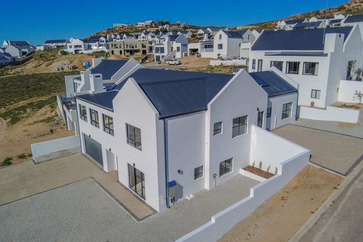 Aerial view of a modern white house with a dark metal roof, featuring a paved driveway and surrounding boundary walls in a coastal estate.
