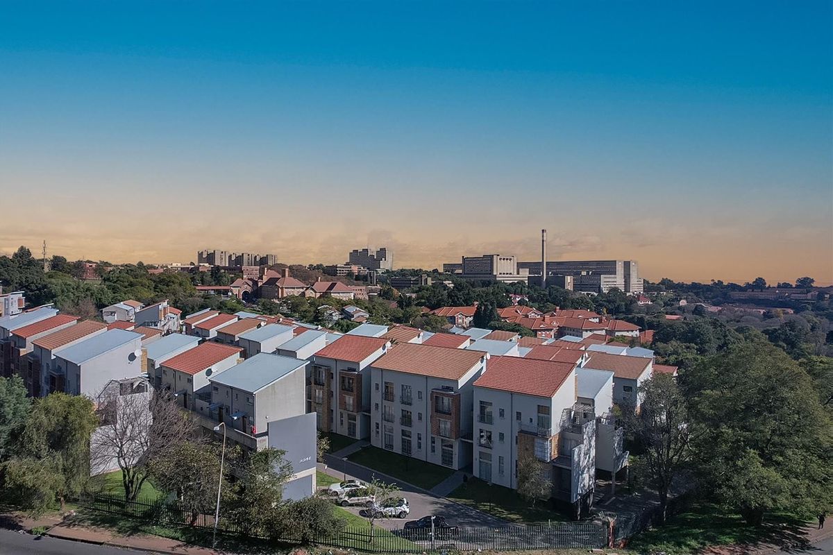 An aerial view of a residential apartment complex featuring multiple buildings with tiled roofs, surrounded by established trees and urban infrastructure.