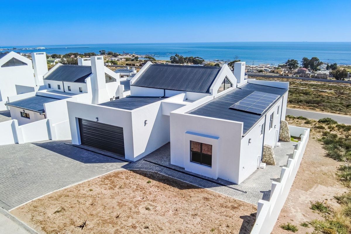 Aerial view of a modern coastal home featuring a dark metal roof, solar panels, white exterior walls, and an attached garage.