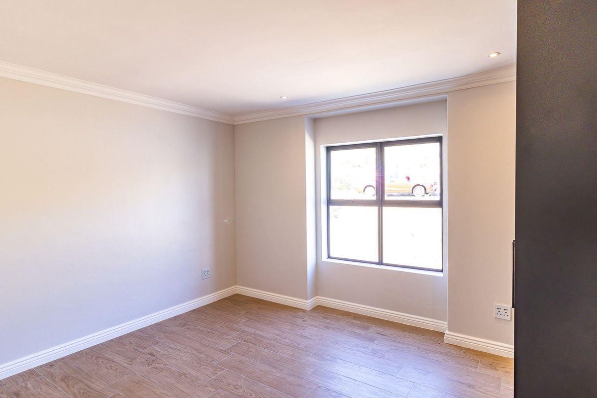A bright, empty bedroom featuring neutral walls, crown molding, and modern wood-look tile flooring with a large window providing natural light.