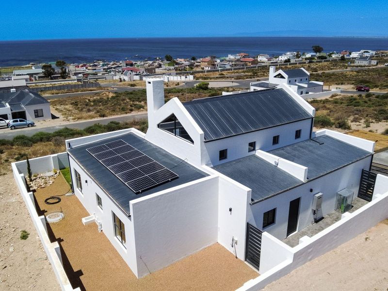 Aerial view of a modern coastal home featuring a white facade, corrugated metal roof, solar panels, and a secure walled perimeter.