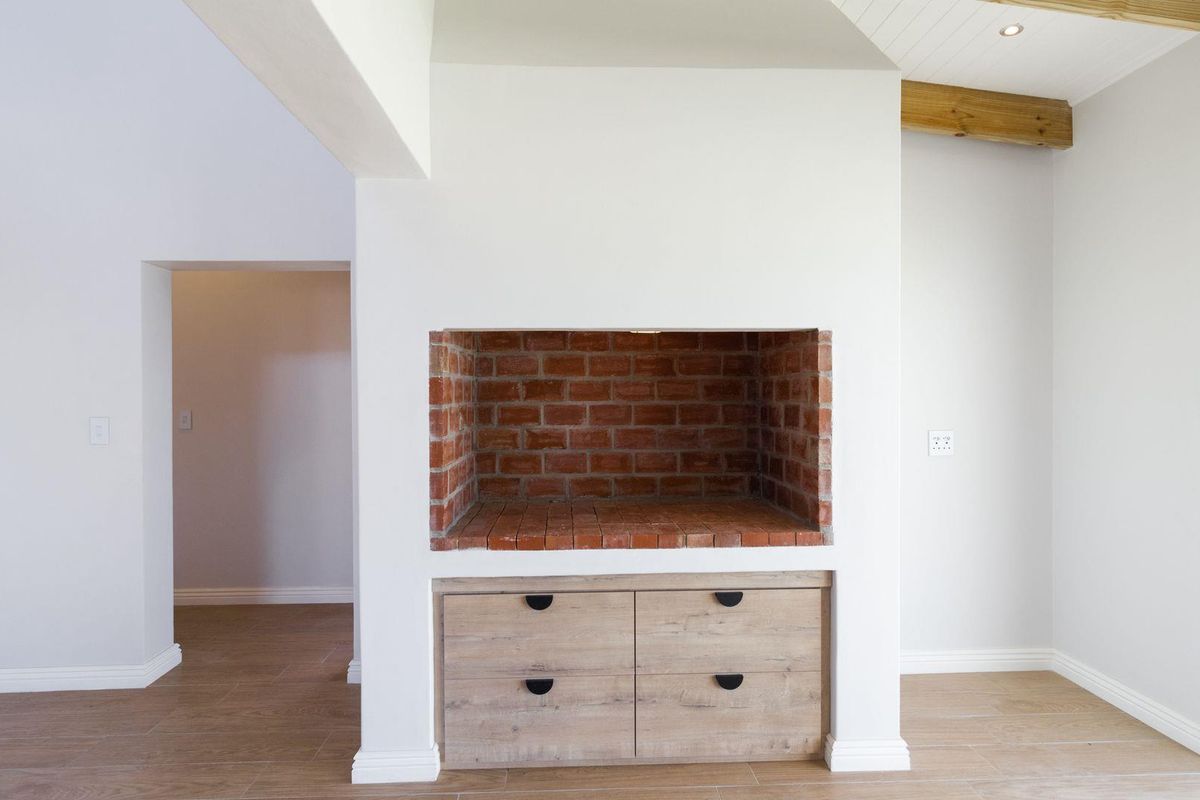 A bright living area featuring a built-in brick fireplace and integrated wooden cabinetry, complemented by modern tiled flooring and exposed ceiling beams.