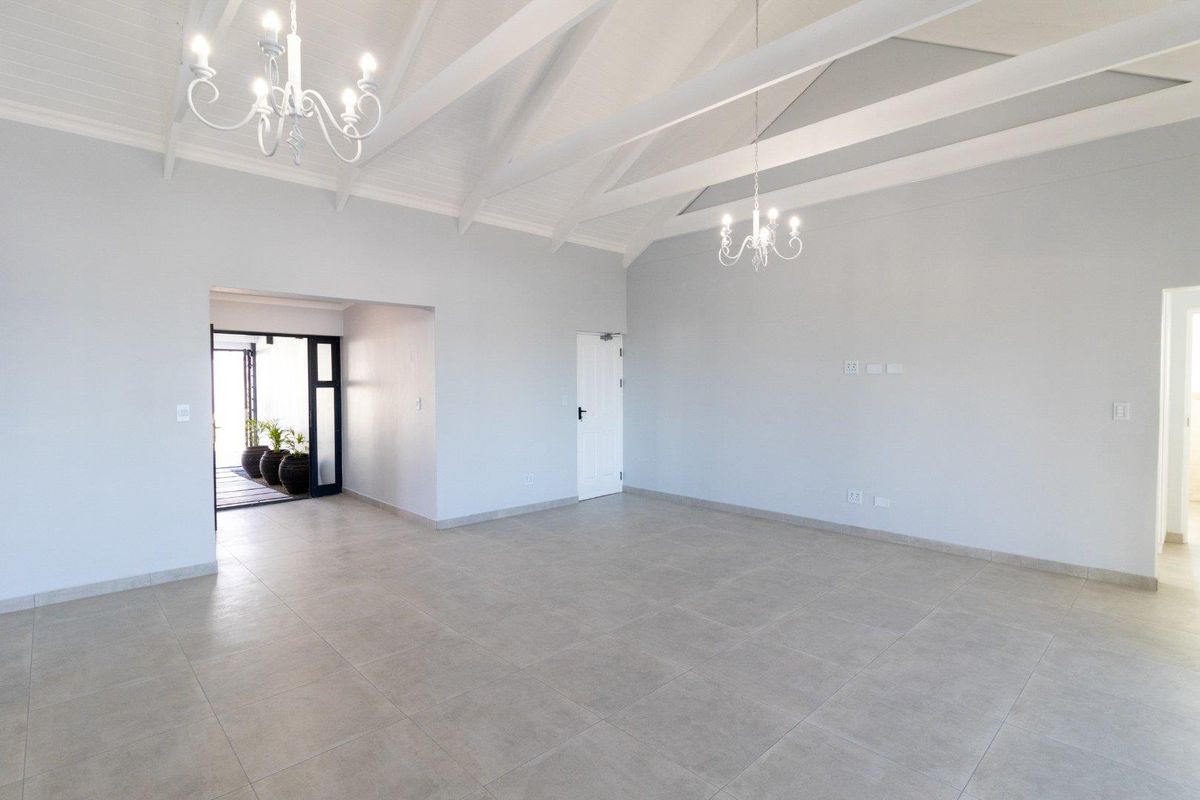 Spacious living area featuring vaulted ceilings with exposed beams, elegant chandeliers, and neutral tiled flooring, leading to an outdoor entrance.
