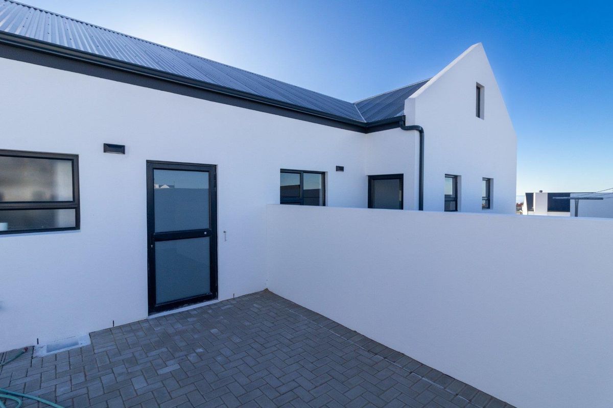 A clean, modern exterior rear view featuring a paved patio area, white rendered walls, dark metal roofing, and black-framed windows and doors.