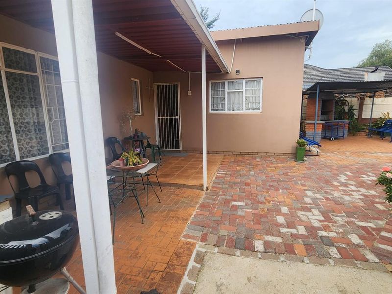 A rear exterior view featuring a covered patio area with tiled flooring, adjacent to a paved courtyard and a built-in brick braai area.