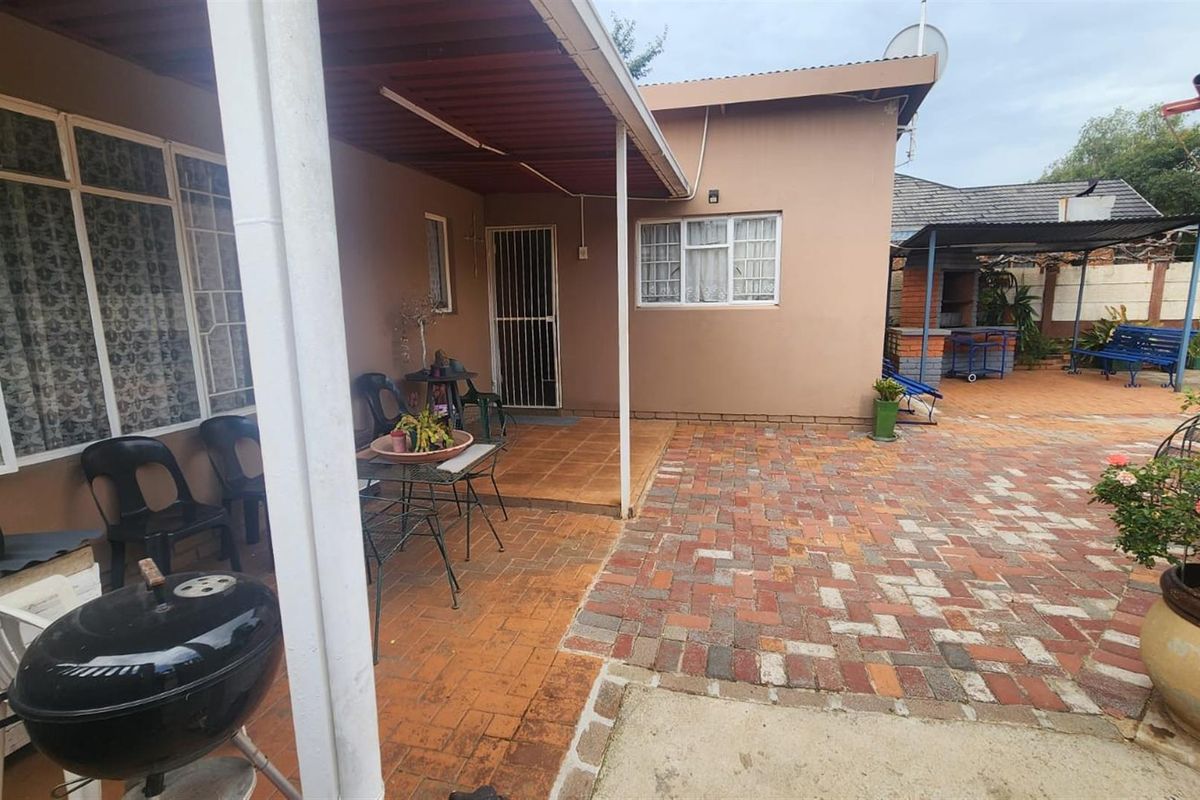 A rear exterior view featuring a covered patio area with tiled flooring, adjacent to a paved courtyard and a built-in brick braai area.