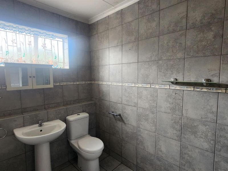 A functional bathroom featuring grey wall tiles, a pedestal basin, a standard toilet, and a window with security bars.