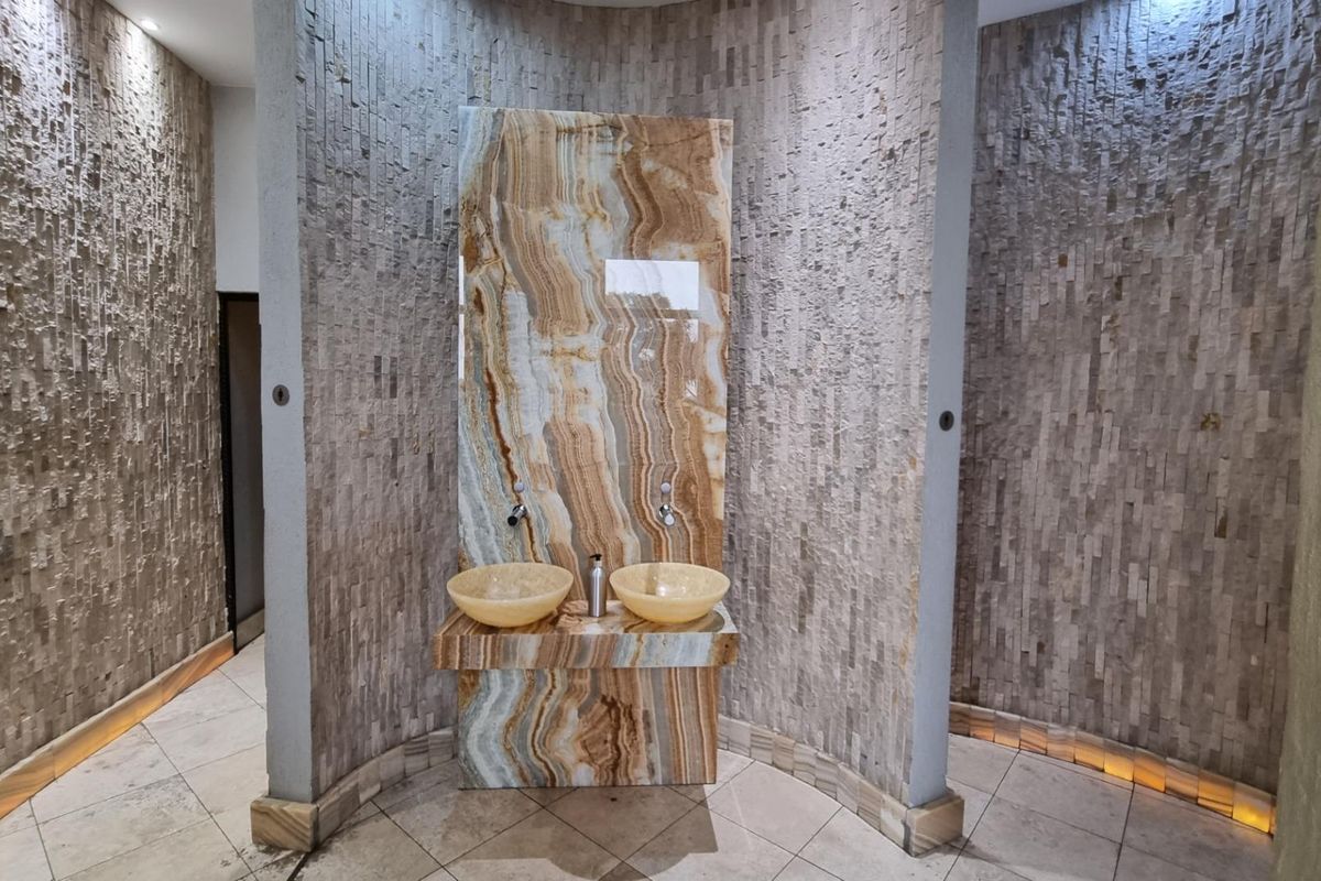 A modern commercial bathroom featuring a striking onyx vanity with dual vessel sinks and textured stone wall cladding with integrated floor lighting.